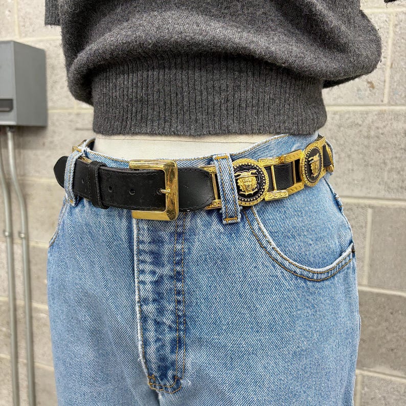 Puede incluir: Un cintur&oacute;n de cuero negro con una hebilla dorada y eslabones met&aacute;licos dorados decorativos con medallones negros circulares que muestran un emblema de cabeza de Medusa dorada en relieve, usado sobre jeans azul claro y un su&eacute;ter de punto gris.