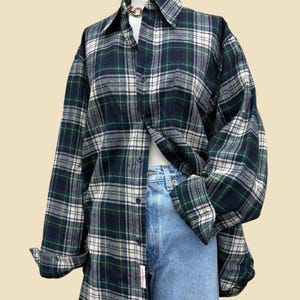 May include: A long-sleeve button-down shirt featuring a dark green, white, and thin yellow plaid pattern. The shirt is worn over light blue denim jeans. The garment has a classic collar, a front button placket, and cuffed sleeves.