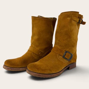 May include: A pair of mid-calf boots crafted from tobacco-colored suede leather. Each boot features a rounded toe, a low stacked heel, and a decorative strap across the vamp secured with a dark metal buckle. An additional strap wraps around the top.