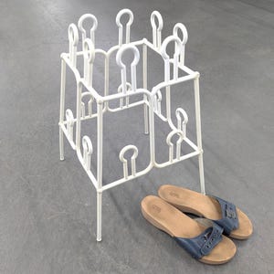 May include: A white metal boot rack with a square frame and multiple vertical prongs featuring circular tops, accompanied by a pair of blue leather sandals with wooden soles and silver buckles positioned on the floor next to the rack.