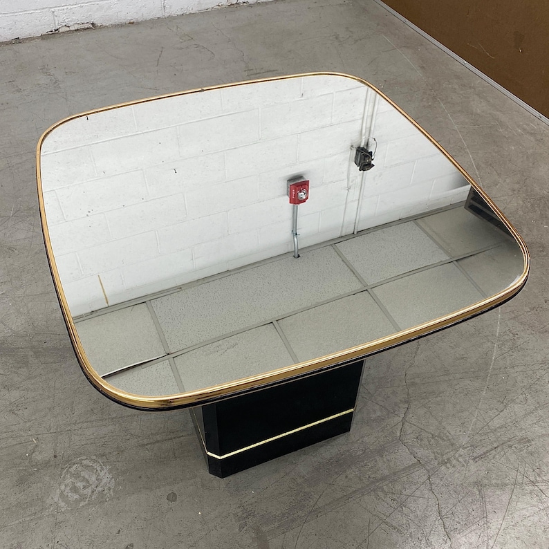 May include: A square coffee table with a mirrored top and a gold-colored trim. The base is black with a gold accent. The table is approximately 30 inches wide and 30 inches long.