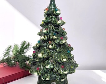 May include: A glossy dark green ceramic Christmas tree figurine with a textured, layered branch design. The tree is decorated with small, multi-coloured translucent plastic bulbs and features a silver metal toggle switch at the base.