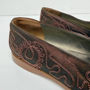 May include: Close-up of a pair of brown satin flats with intricate embroidered detailing in a darker brown hue. The shoes have a low heel and a leather sole. The embroidery features swirling patterns and floral motifs.