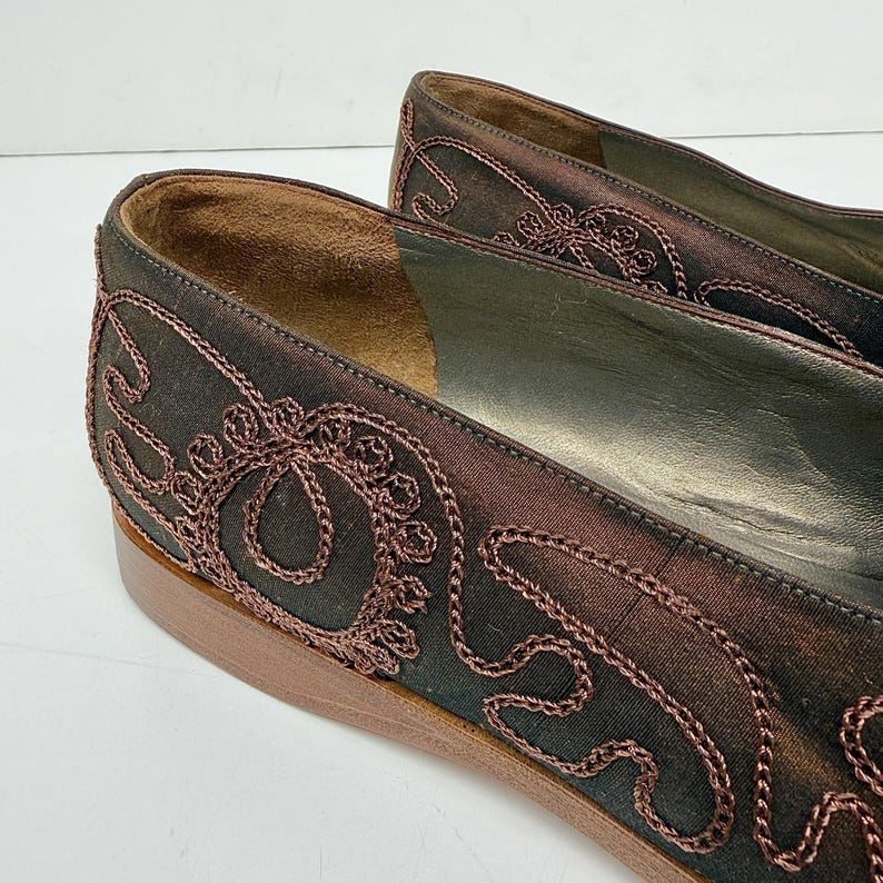 May include: Close-up of a pair of brown satin flats with intricate embroidered detailing in a darker brown hue. The shoes have a low heel and a leather sole. The embroidery features swirling patterns and floral motifs.