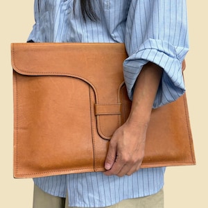 May include: A tan leather document portfolio featuring a large flap closure with a vertical strap and a single loop fastener. The portfolio has a smooth, matte finish and visible stitching along the edges, providing a structured, flat design.