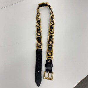 Puede incluir: Un cintur&oacute;n de cuero negro que presenta una serie de medallones circulares de metal dorado con dise&ntilde;os en relieve. El cintur&oacute;n incluye una hebilla rectangular de metal dorado y una trabilla a juego, con un extremo puntiagudo con cinco agujeros.