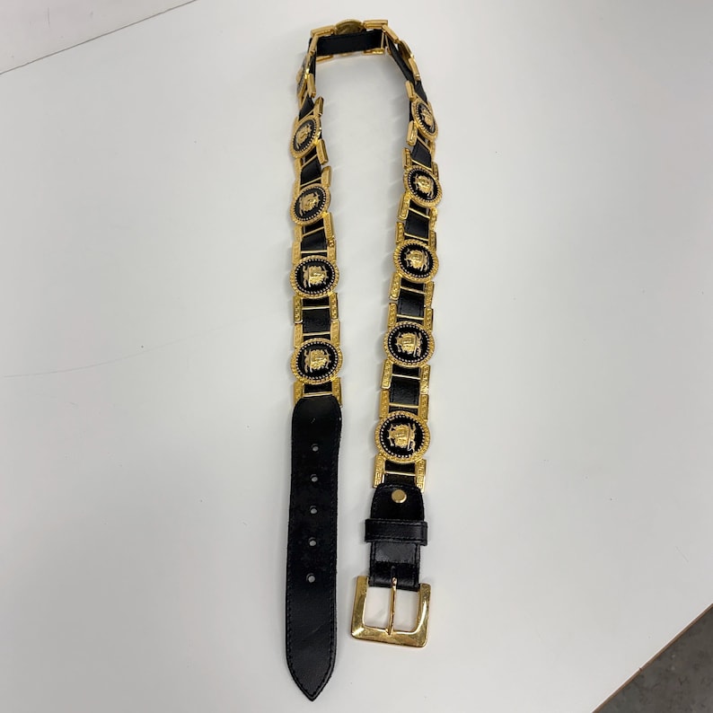 Puede incluir: Un cintur&oacute;n de cuero negro que presenta una serie de medallones circulares de metal dorado con dise&ntilde;os en relieve. El cintur&oacute;n incluye una hebilla rectangular de metal dorado y una trabilla a juego, con un extremo puntiagudo con cinco agujeros.