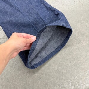 May include: Close-up of blue denim trousers. The fabric is a medium wash denim with visible texture. The hem of the trousers is turned up, revealing the inside seam. The trousers are on a grey surface.