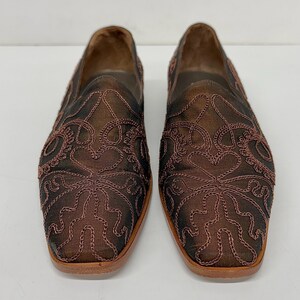 May include: A pair of brown slip-on shoes with detailed embroidered floral patterns. The shoes feature a square toe and a light brown sole. The upper material is a dark brown fabric with lighter brown embroidery.
