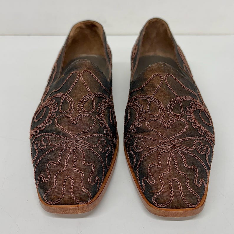 May include: A pair of brown slip-on shoes with detailed embroidered floral patterns. The shoes feature a square toe and a light brown sole. The upper material is a dark brown fabric with lighter brown embroidery.