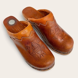 May include: A pair of tan leather clogs featuring a rounded toe, a slip-on design, and a decorative embossed geometric pattern on the upper. The shoes have a visible stitched zigzag detail along the collar and are secured with small metal studs.