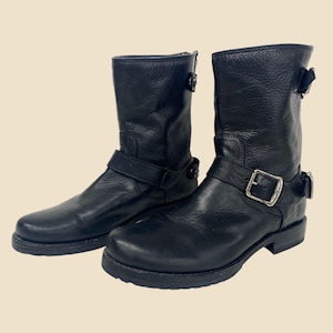 May include: A pair of black leather motorcycle boots. Each boot features a buckle strap and a short shaft. The boots have a textured appearance and a rounded toe. The soles are black.