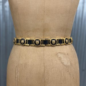 Puede incluir: Un cintur&oacute;n que presenta segmentos de cuero negro alternados con medallones circulares de metal dorado. Cada medall&oacute;n muestra un motivo de cabeza en relieve sobre un fondo de esmalte negro, unidos por eslabones rectangulares dorados.