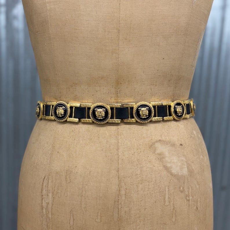 Puede incluir: Un cintur&oacute;n que presenta segmentos de cuero negro alternados con medallones circulares de metal dorado. Cada medall&oacute;n muestra un motivo de cabeza en relieve sobre un fondo de esmalte negro, unidos por eslabones rectangulares dorados.