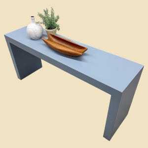 May include: A modern, light blue console table with a minimalist design. A wooden bowl, a white vase with a black design, and a potted plant sit on the table. The table's legs are angled, creating a unique silhouette.