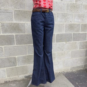 May include: Dark blue denim bell-bottom trousers with a brown leather belt and gold buckle. The trousers have a classic five-pocket design and a flared leg. The model is wearing a pink top with a red apple pattern.