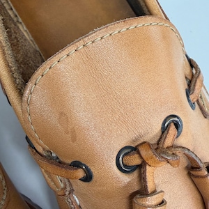 May include: A tan leather boat shoe featuring a moccasin-style toe construction with visible light-colored stitching along the vamp. The shoe includes dark metal eyelets and matching tan leather laces threaded through the sides and tied at the front.