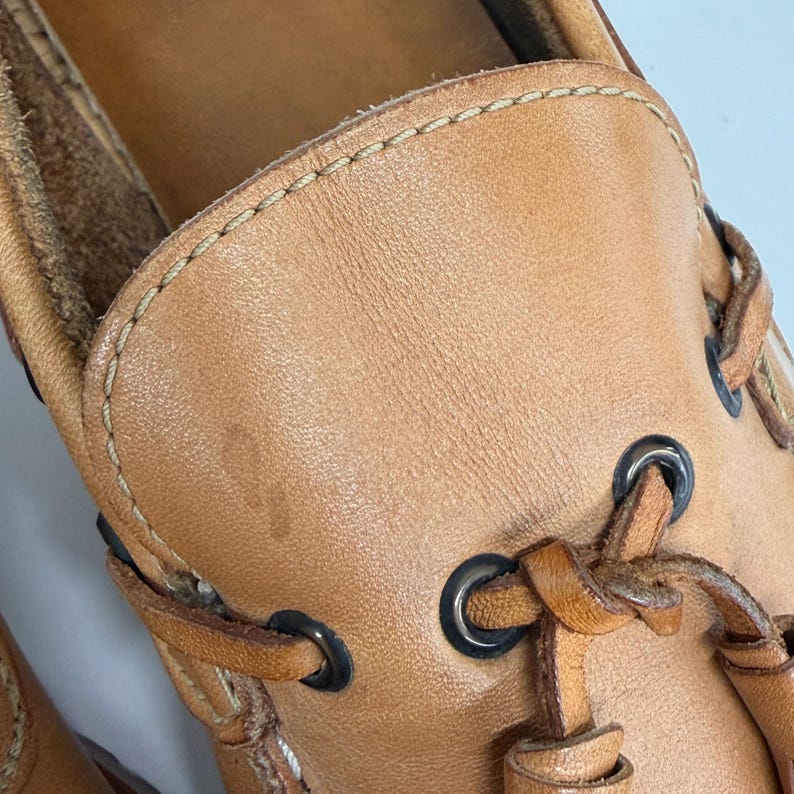 May include: A tan leather boat shoe featuring a moccasin-style toe construction with visible light-colored stitching along the vamp. The shoe includes dark metal eyelets and matching tan leather laces threaded through the sides and tied at the front.