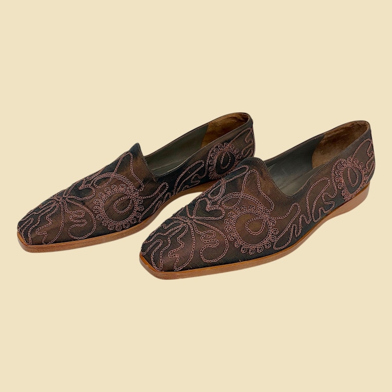 May include: A pair of brown slip-on shoes with detailed embroidered floral patterns. The shoes feature a square toe and a low heel. The embroidery is a darker brown, contrasting with the lighter brown base material.