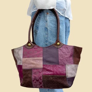 May include: A multi-textured patchwork tote bag featuring various shades of purple, pink, and brown. The bag is constructed from panels of leather, suede, and fabric with a repeating embossed logo pattern, accented by dark brown leather handles and brass hardware.
