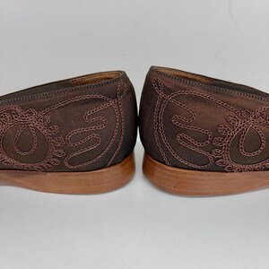 May include: A pair of brown satin flats with embroidered detailing. The shoes have a low heel and a decorative stitched design on the sides. The soles are a light brown colour, and the shoes are set against a white background.