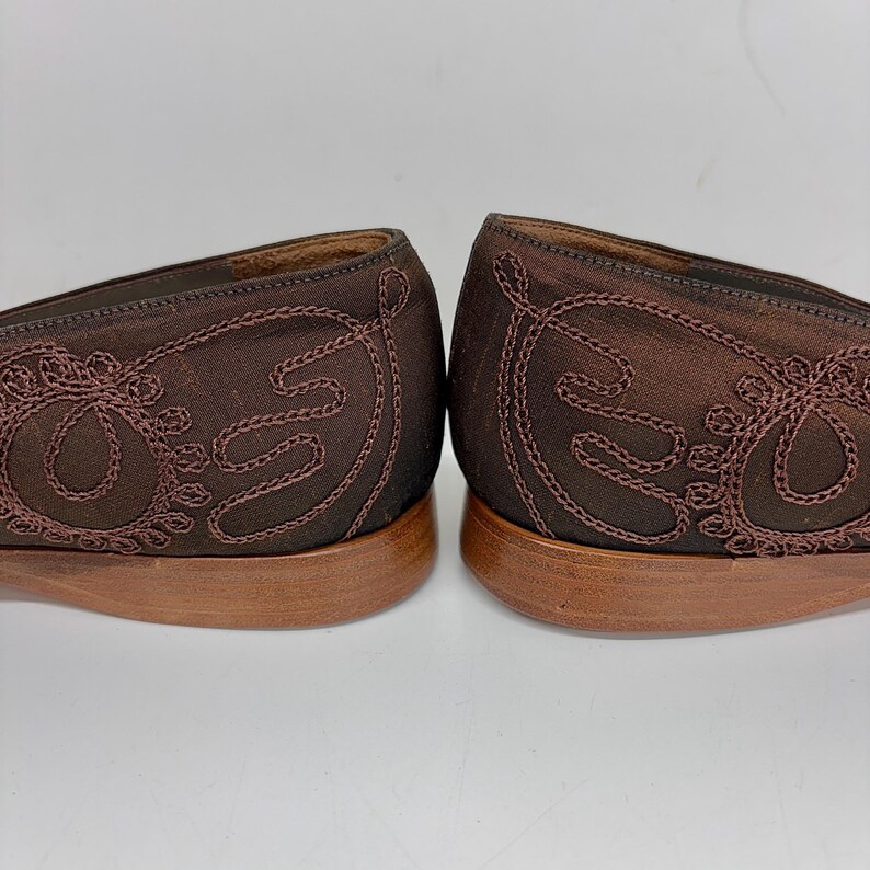 May include: A pair of brown satin flats with embroidered detailing. The shoes have a low heel and a decorative stitched design on the sides. The soles are a light brown colour, and the shoes are set against a white background.