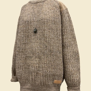 May include: A long-sleeve, ribbed knit sweater in a heathered brown and tan yarn. The garment features solid tan shoulder patches, a crew neckline, and a small rectangular tan leather-like brand patch on the lower left side.