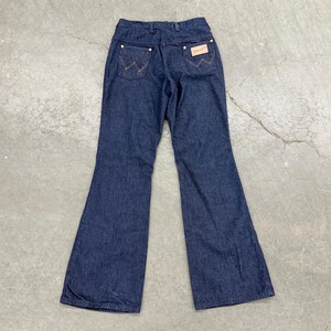 May include: Dark blue denim Wrangler jeans with a flared leg. The back pockets feature the signature Wrangler stitching in a contrasting tan thread. A tan leather patch with the Wrangler logo is visible.