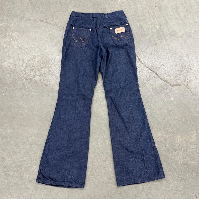 May include: Dark blue denim Wrangler jeans with a flared leg. The back pockets feature the signature Wrangler stitching in a contrasting tan thread. A tan leather patch with the Wrangler logo is visible.