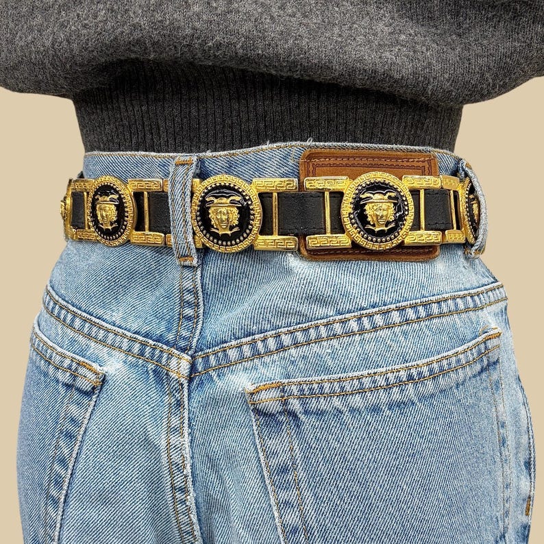 Puede incluir: Un cintur&oacute;n de cuero negro con herrajes de metal dorado y medallones circulares que muestran un motivo de rostro en relieve. El cintur&oacute;n incluye eslabones rectangulares con un patr&oacute;n de greca y una hebilla rectangular de cuero marr&oacute;n.