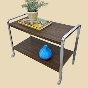 May include: A two-tiered serving cart with a dark wood grain finish and chrome-colored metal frame. The top shelf holds a small potted plant and a vintage book. The bottom shelf displays a blue and purple striped glass vase. The cart has small wheels.