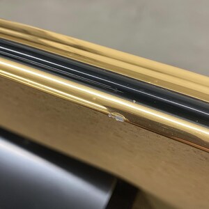 May include: Close-up of a gold-colored frame with a black stripe. The frame has a shiny, reflective surface. The image is angled, showing the corner of the frame.