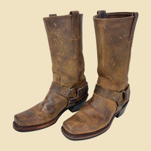 May include: A pair of brown leather boots with a distressed finish. The boots have a square toe, a harness strap around the ankle, and a pull tab at the top. The boots are approximately 12 inches tall.