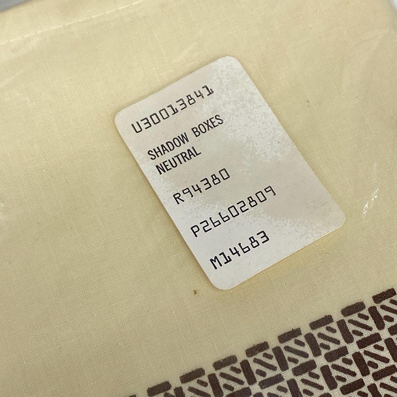 May include: A close-up shot of a cream-colored fabric with a printed brown geometric pattern along the bottom edge. A white rectangular tag is placed on the fabric, with the text "SHADOW BOXES NEUTRAL" and other numerical codes.