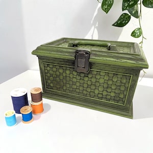 May include: A rectangular olive green storage box with a textured basket-weave pattern and a dark metal latch. Beside the box are four small spools of thread in blue, light blue, orange, and brown, each with a white plastic center core.