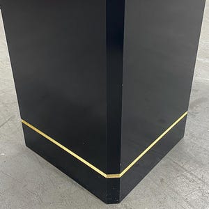 May include: A black, rectangular pedestal with a glossy finish. The edges are accented with a thin, gold-colored trim. The pedestal is a solid, geometric shape, suitable for display.