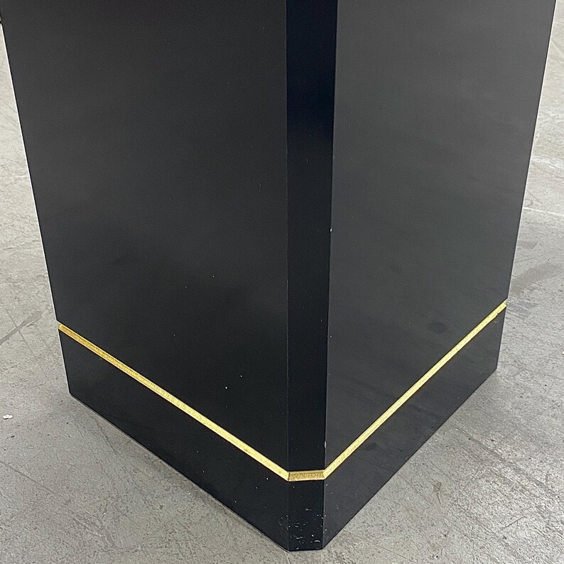May include: A black, rectangular pedestal with a glossy finish. The edges are accented with a thin, gold-colored trim. The pedestal is a solid, geometric shape, suitable for display.