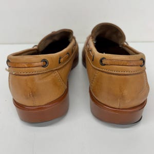 May include: A pair of tan leather loafers featuring a moccasin-style construction with a decorative lace detail threaded through dark metal eyelets around the collar and a solid, smooth brown heel and sole base for a classic footwear appearance.