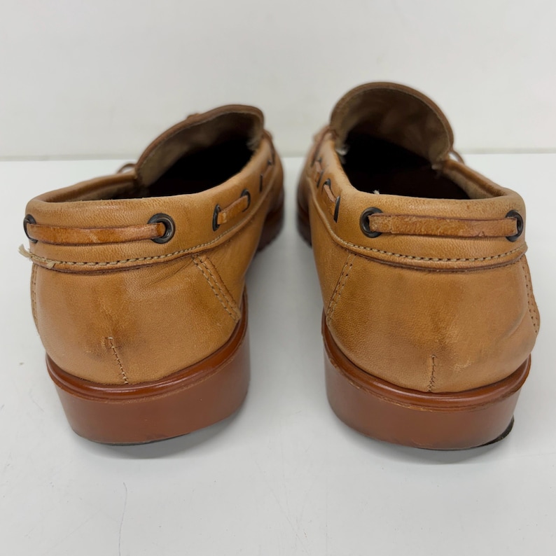 May include: A pair of tan leather loafers featuring a moccasin-style construction with a decorative lace detail threaded through dark metal eyelets around the collar and a solid, smooth brown heel and sole base for a classic footwear appearance.
