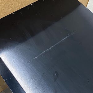May include: A close-up of a black, glossy surface with a long, light-colored scratch. The surface appears to be part of a larger object, possibly a table or a piece of furniture. The black finish reflects light, highlighting the scratch.