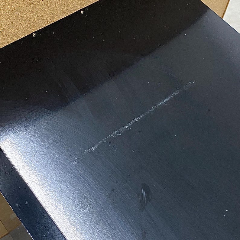 May include: A close-up of a black, glossy surface with a long, light-colored scratch. The surface appears to be part of a larger object, possibly a table or a piece of furniture. The black finish reflects light, highlighting the scratch.