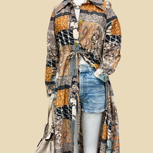 May include: A long-sleeved, floor-length duster coat featuring a multi-colored patchwork pattern in shades of mustard yellow, black, white, and blue. The coat has a collared neckline, front button closures, and a tie waist, worn over light blue denim shorts.