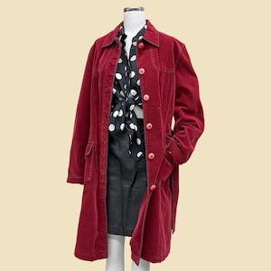 May include: A long-sleeved, deep red coat featuring a collared neckline, a front button closure with matching red buttons, and two front pockets. Underneath, a black shirt with a white polka dot pattern is tied at the waist over a black skirt.