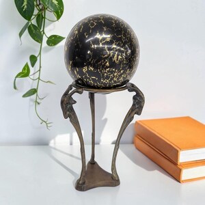 May include: A smooth, spherical object with a black surface featuring intricate gold-colored veining patterns. The sphere rests securely on a tripod-style metal stand with a dark, weathered bronze finish and three curved, slender legs.