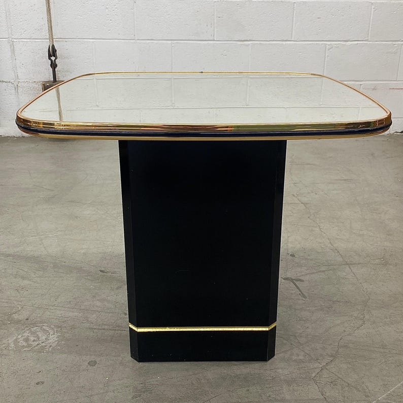 May include: A black side table with a mirrored top and gold trim. The table has a rectangular base and a square top. The table is approximately 24 inches tall and 24 inches wide.