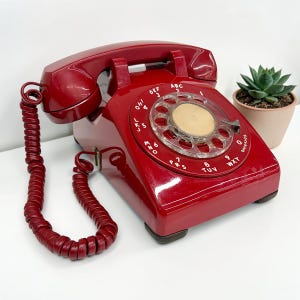 May include: A vintage red rotary dial telephone with a matching coiled handset cord. The phone features a clear dial with numbers and letters, and a glossy finish. A small succulent plant in a terracotta pot is in the background.