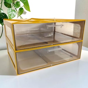 May include: A stackable storage unit featuring two rectangular drawers with mustard yellow frames and clear plastic fronts. Each drawer includes a small, horizontal, transparent handle centered on the front panel for easy access to the interior.