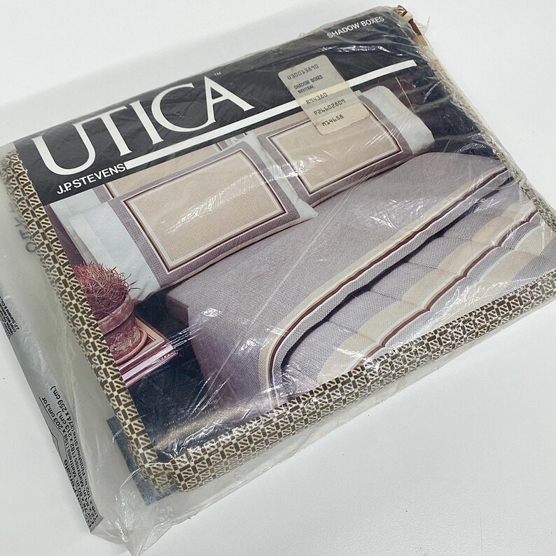 May include: A packaged set of Utica brand shadow boxes bedding. The bedding set includes pillowcases and a comforter with a neutral color palette of beige, gray, and brown. The packaging is clear plastic, and the brand name is prominently displayed.