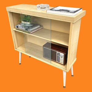 May include: A vintage wooden cabinet with glass sliding doors, featuring a small radio, books, and a plant. The cabinet has white legs and a light wood finish. An ashtray and a book are on top.