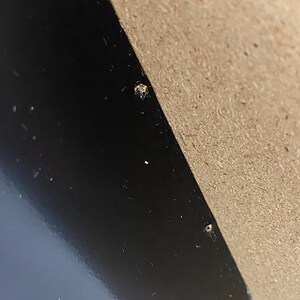 May include: A close-up shot of a black, glossy surface with a diagonal section of light brown cardboard. Small, light-colored specks are scattered across the black surface. The cardboard has a rough texture.